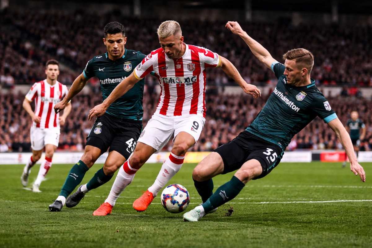 Dynamic moment from a Stoke City vs Southampton match showing a Southampton defender trying to intercept a Stoke City forward on the pitch, with fans cheering in the background at bet365 Stadium.