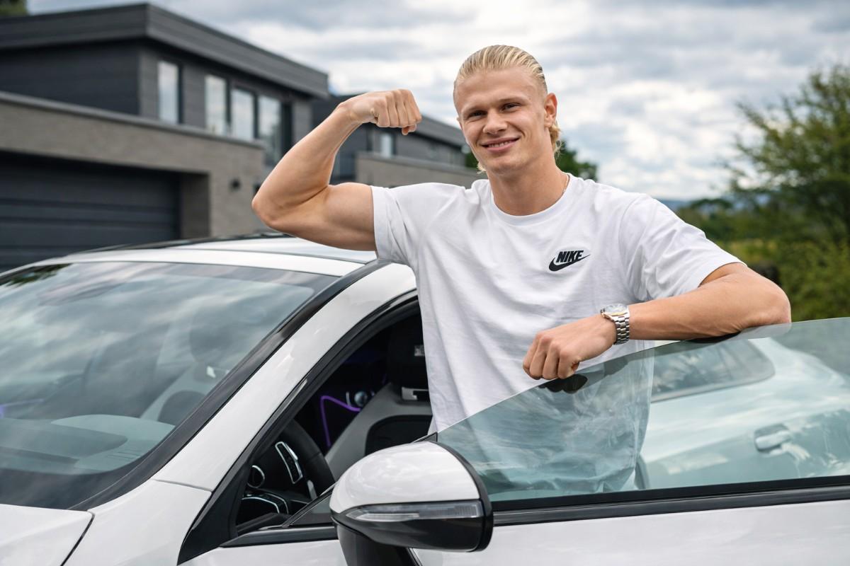 Erling Haaland posing beside a sports car while flexing his arm.